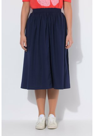 Person, die einen marineblauen Midirock, ein rotes Oberteil und weiße Turnschuhe trägt, steht vor einem schlichten weißen Hintergrund.