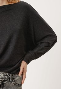 Svart, löst sittande tröja med en mjuk textur och subtil lyster. Har vida ärmar och båtformad halsringning. Matchas med grå jeans.