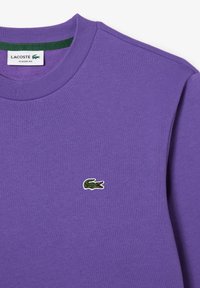 Lilla crewneck sweatshirt med lille grøn broderet Lacoste krokodille logo på venstre bryst og hvidt label med teksten "Lacoste Classic Fit" indeni kraven.