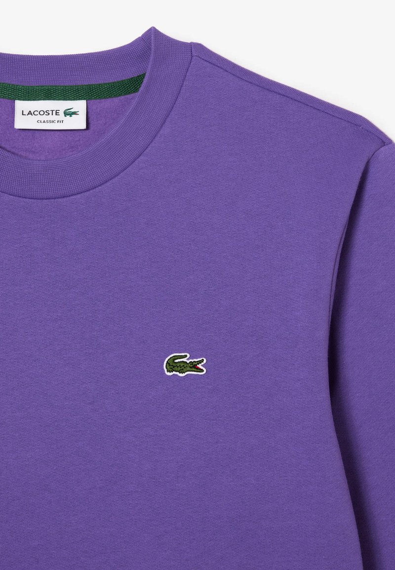Fioletowa bluza crewneck z małym, zielonym, wyhaftowanym logo krokodyla Lacoste na lewej piersi oraz białą metką z napisem "Lacoste Classic Fit" wewnątrz kołnierza.