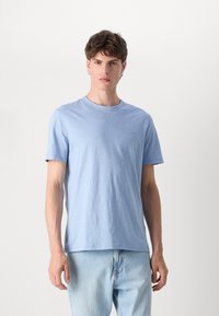 T-shirt azul claro de manga curta em tecido suave e texturizado, com decote redondo e um logo discreto no peito. Usado com jeans de denim claros.