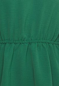 Tessuto verde con una superficie liscia e testurizzata, caratterizzato da una vita elastica arricciata. Il materiale ha una leggera lucentezza ed è leggero.