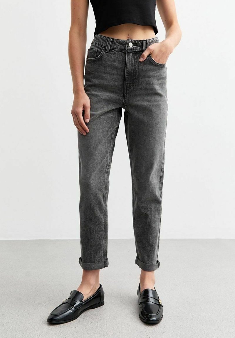 Personne portant un jean gris foncé retroussé, un top court noir et des mocassins en cuir noir, debout sur un sol gris face à un mur blanc.