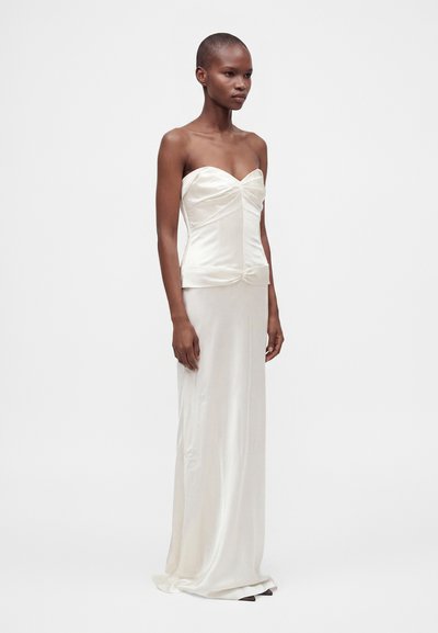 Femme aux cheveux courts portant une robe blanche soyeuse bustier, debout devant un fond blanc uni.