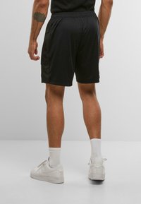 Man standing wearing black athletic shorts, white socks, and white Nike sneakers, with a leaf tattoo visible on left forearm.