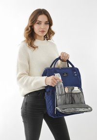 Marinblå ryggsäck med kviltad textur, två handtag på toppen och ett framfack som innehåller en flaska, sked och behållare.