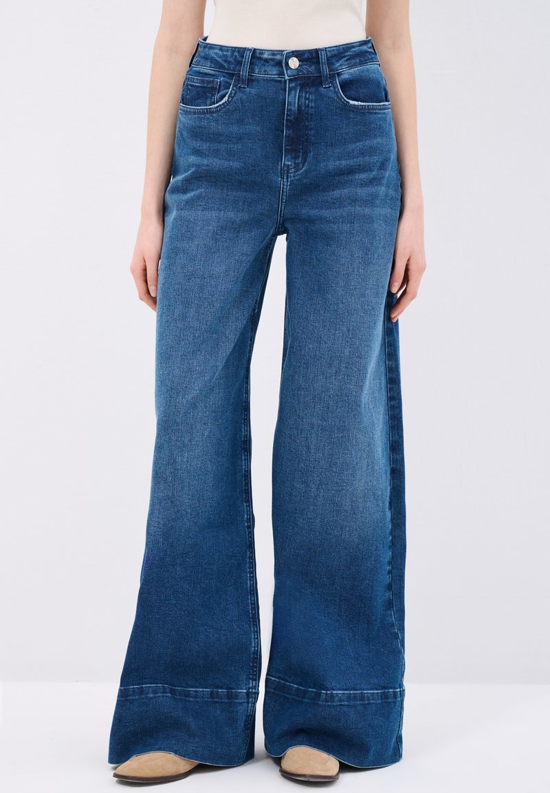 Jeans de mezclilla azul de tiro alto con un diseño de pierna ancha, que cuentan con una cremallera de botones, dos bolsillos frontales y un ligero desgaste en la tela.