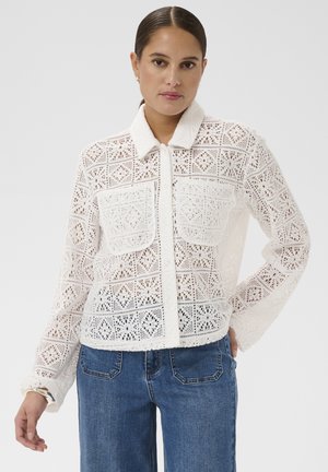 Femme portant une chemise blanche en crochet à manches longues avec deux poches avant et un jean en denim bleu, posant avec une main sur la hanche.
