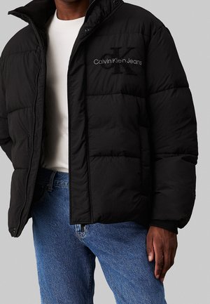 Mężczyzna ubrany w czarną kurtkę puchową Calvin Klein Jeans, białą koszulę i niebieskie dżinsy, z ręką swobodnie opuszczoną wzdłuż ciała.