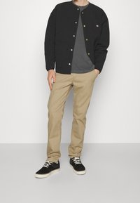 Czarna kurtka zapinana na guziki z okrągłym dekoltem, khaki spodnie i czarne buty z białymi podeszwami. Gładki materiał, casualowy design i widoczne przeszycia.