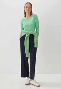 Groene langbouwtop gecombineerd met marineblauwe broek met wijde pijpen, aangevuld met een lichtgroene gebreide trui die op de taille is geknoopt en witte sneakers.