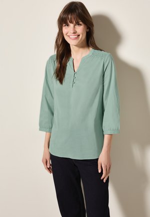 Blusa verde claro de tres cuartos con escote en V, con detalles plisados cerca de los hombros y una tapeta de tres botones.