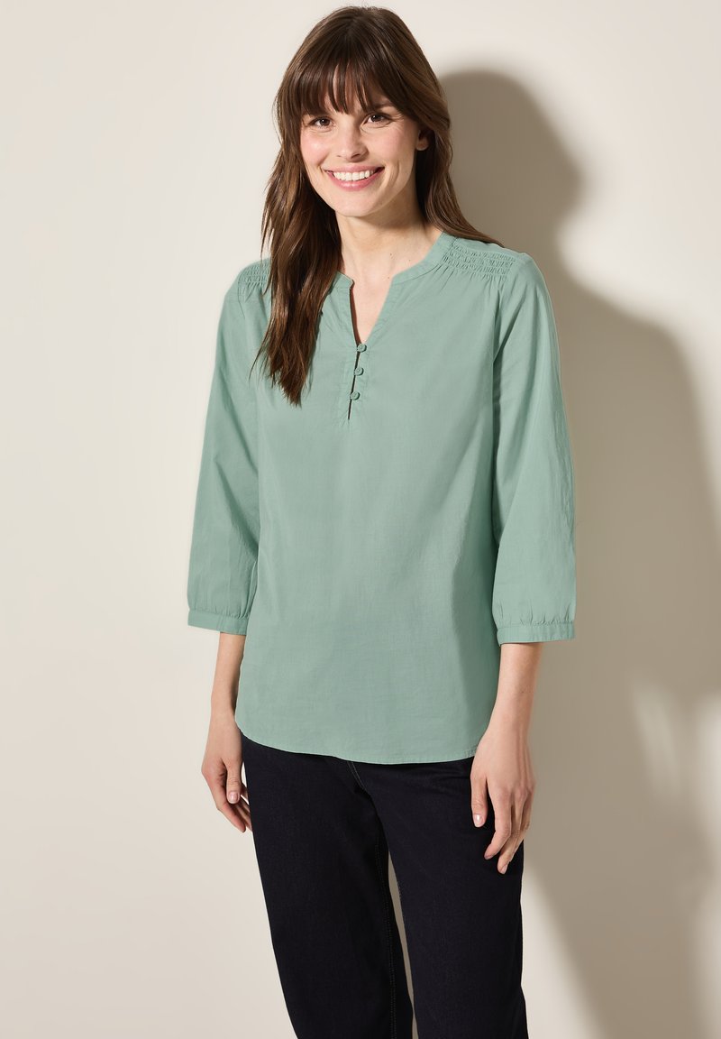 Blusa verde chiaro con maniche a tre quarti e scollo a V, caratterizzata da dettagli plissettati vicino alle spalle e una chiusura con tre bottoni.