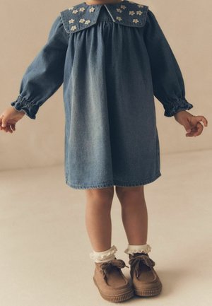 Robe en denim avec manches bouffantes et grand col, ornée de broderies florales. Chaussures marron à finition texturée et chaussettes blanches froncées.