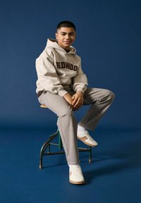 Cremefarbener Hoodie mit "REDWOOD"-Aufdruck, graue Hose und weiße Sneakers mit braunen Akzenten. Sitzt auf einem grünen Hocker vor einem dunkelblauen Hintergrund.