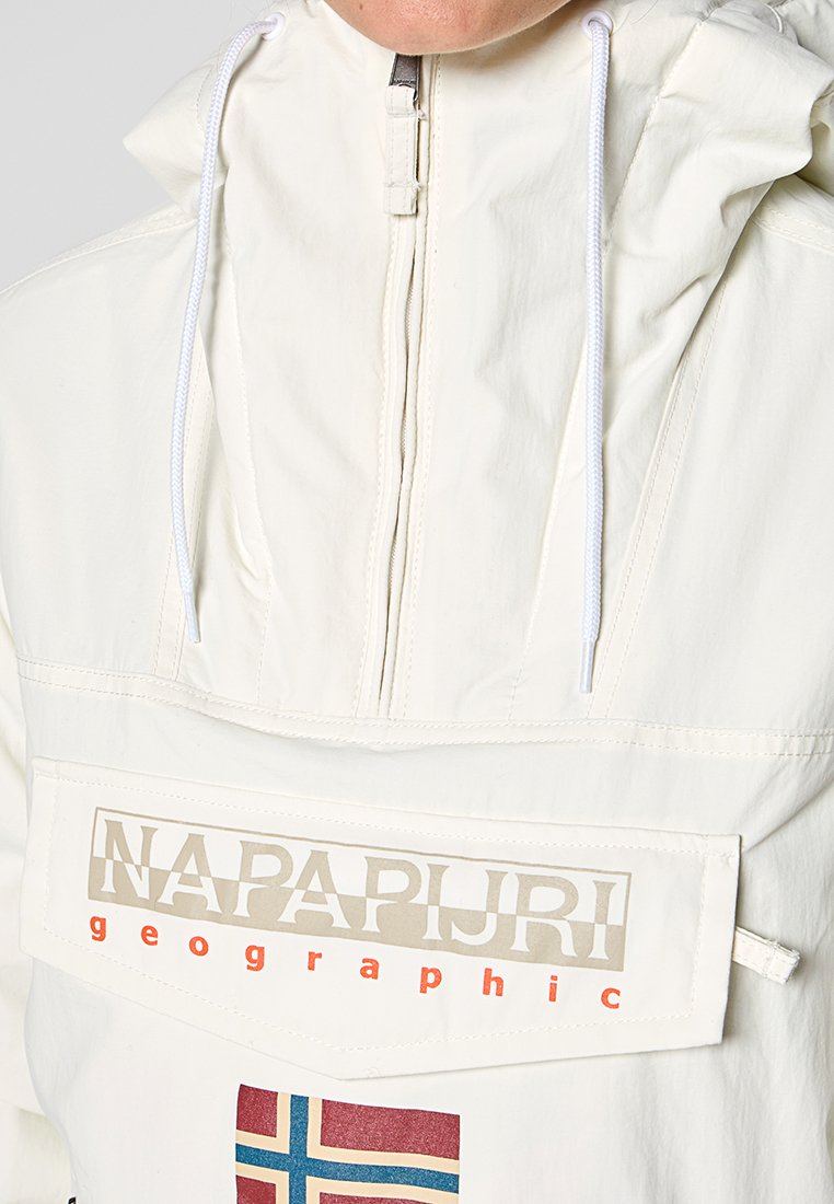 Eine Person trägt eine cremefarbene Kapuzenjacke mit einer Fronttasche, die das "Napapijri geographic"-Logo und ein kleines norwegisches Flaggenpatch darunter zeigt.
