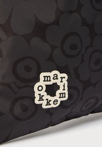 Μαύρο ύφασμα με σκοτεινό ανθικό σχέδιο και λευκή λαστιχένια πινακίδα logo που γράφει «marimekko» σε κυκλική διάταξη.