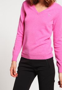 Pull rose en V fabriqué à partir d'un matériau doux, avec des manches longues et des poignets côtelés. Petit logo sur la poitrine. Associé à un pantalon noir.