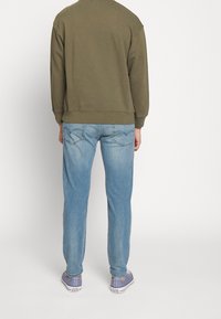 Olivově zelená mikina, světle modré džínové kalhoty a šedé boty. Mikina má žebrované manžety a lem; džíny mají detaily prošívání na zadních kapsách.