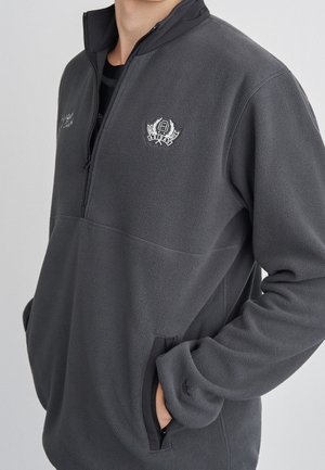 Mann trägt dunkelgrauen Viertel-Reißverschluss-Fleecepullover mit gesticktem Logo auf der Brust und Hand in Reißverschlusstasche an der Seite vor einfarbigem Hintergrund.