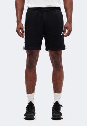Homme portant un short Adidas noir avec des bandes blanches sur les côtés, des chaussettes blanches mi-mollet et des chaussures de course noires, debout les mains détendues.