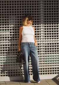 Valkoinen ribattu hihaton paita, yhdistettynä löysiin sinisiin farkkuihin, pidetään mustaa käsilaukkua taustalla, jossa on ruudukkoasu.