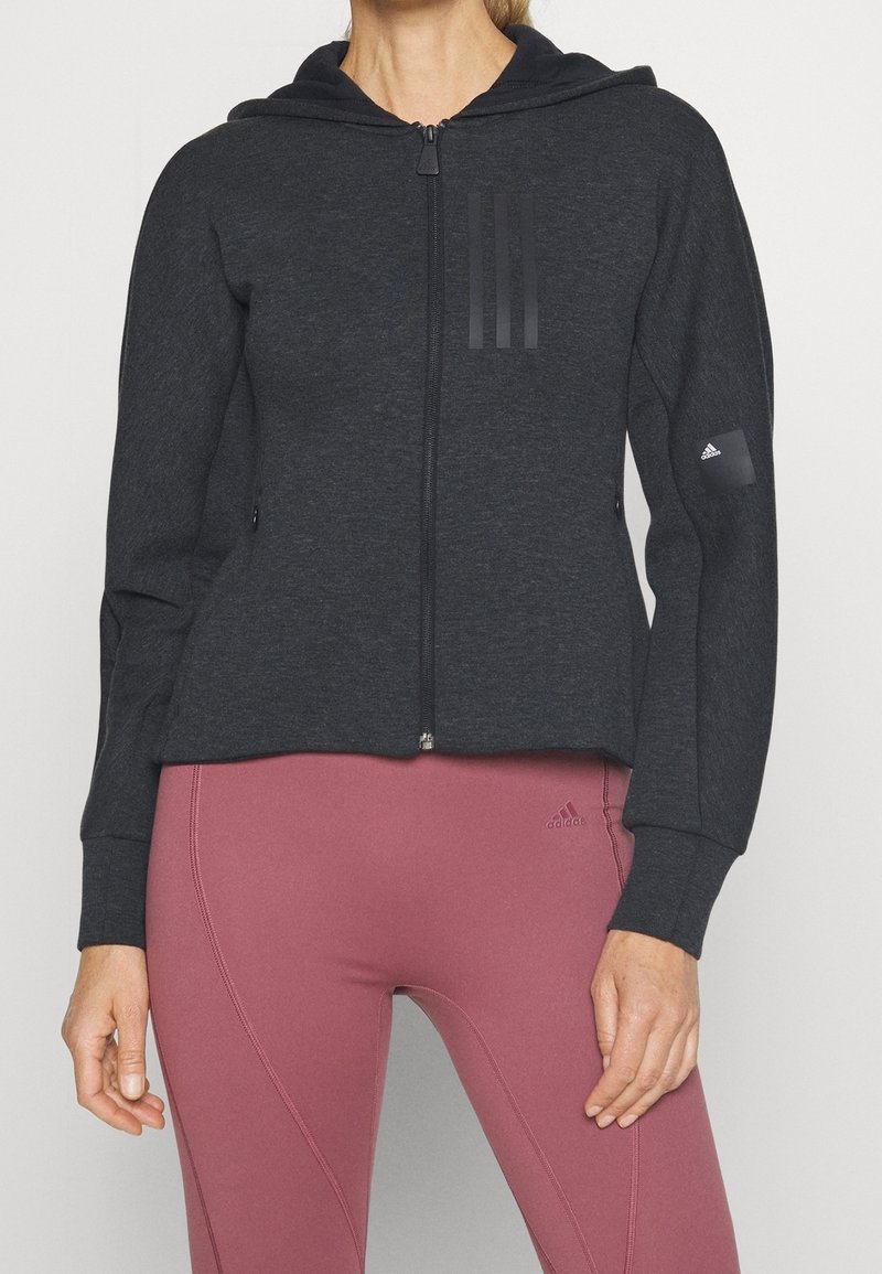 Haut à capuche zippé gris foncé en matière douce, avec trois rayures en relief sur la poitrine et une étiquette de logo sur la manche droite.