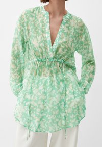Donna che indossa una blusa floreale verde menta trasparente con maniche lunghe e vita regolabile, abbinata a pantaloni bianchi.