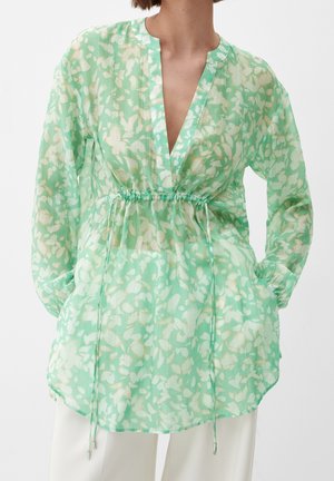 Kvinna i en genomskinlig mintgrön blommig blus med långa ärmar och dragsko i midjan, paired med vita byxor.
