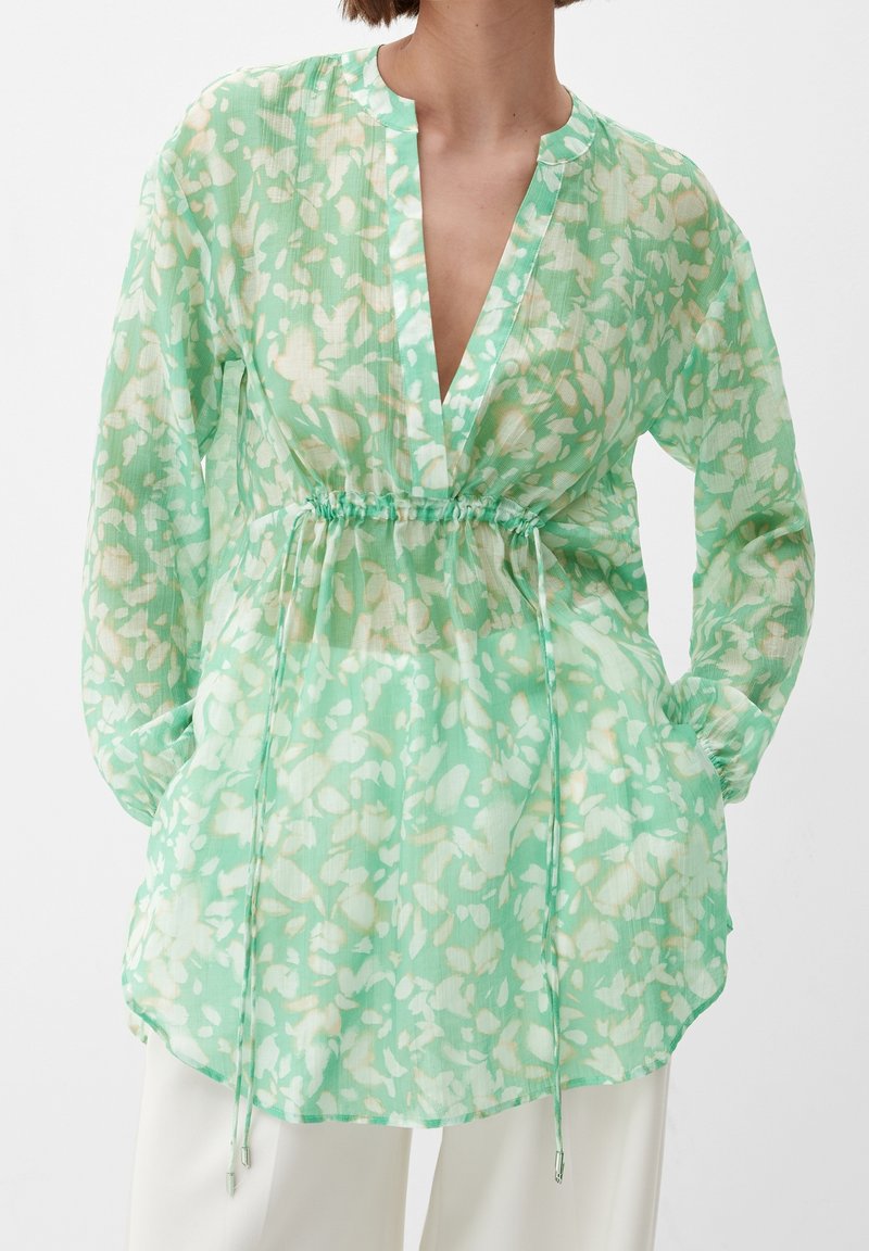 Donna che indossa una blusa floreale verde menta trasparente con maniche lunghe e vita regolabile, abbinata a pantaloni bianchi.