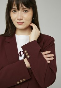 Blazer bordeaux avec un revers cranté, présentant quatre boutons dorés sur les poignets. Superposé à un T-shirt blanc avec un texte bordeaux.
