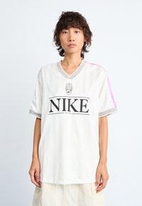Jersey blanco de Nike con rayas verticales, mangas cortas, cuello en V gris y acentos rosas en los hombros; presenta un logotipo negro en bold en el centro.