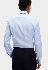 Mężczyzna w jasnoniebieskiej koszuli i ciemnych spodniach w prążki, pokazany z tyłu na jednolitym jasnym tle.