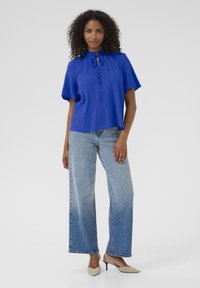 Haut texturé bleu à manches courtes avec un détail à nouer au cou, associé à un jean bleu clair à jams larges et des escarpins beiges pointus.