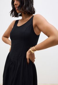 Zwarte mouwloze jurk met een aansluitend lijfje, wijde rok en geplooemde zoom. Soepele textuur met subtiele stikseldetails.