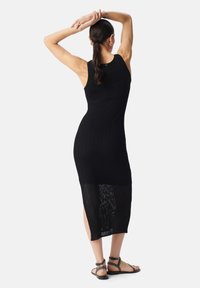Abito in maglia nera a coste con silhouette aderente, lunghezza al ginocchio e una parte inferiore trasparente. Indossato con sandali neri con cinturini.