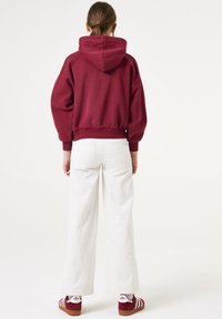 Bordeaux cropped hoodie met een voorste kangaroo pocket, gecombineerd met witte wijde broekspijpen en rode sneakers met witte accenten.