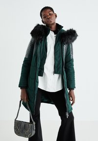 Zaļš, pildīts mētelis ar melnu mākslīgā kažoka kapuci, sudraba panelu akcentiem un priekšējo rāvējslēdzēju; modelis tur pelēku rokas somu ar kniedēm.