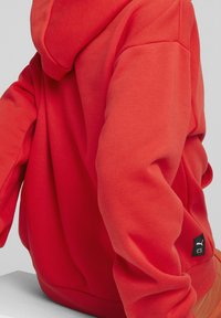 Červená mikina s kapucí vyrobená z měkké látky, se stahovací šňůrkou, žebrovanými manžetami a malou černou nášivkou na dolní straně.