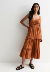 Robe maxi en tissu léger de couleur rouille, à plusieurs niveaux, avec des accents en volants ; associée à des sandales plates noires à glissière avec une finition en boucle.