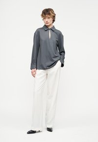 Άτομο στέκεται φορώντας μια μακρυμάνικη μπλούζα σκούρου γκρι με άνοιγμα στο ντεκολτέ και λευκό παντελόνι φαρδιάς γραμμής με μαύρα μυτερά παπούτσια.