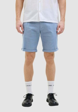 Uomo che indossa pantaloncini azzurri risvoltati, calzini bianchi con scritte nere, scarpe nere con lacci e una camicia bianca a maniche corte, in piedi.