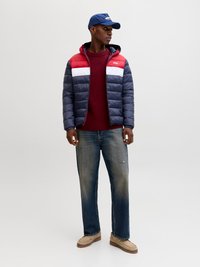 Gevoerde jas met rode, witte en marineblauwe secties, gecombineerd met een bordeauxrode trui, vervaagde blauwe jeans en beige schoeisel. Blauwe pet inbegrepen.