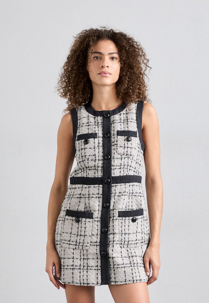 Robe en tweed présentant un motif à carreaux gris et blanc, avec des finitions noires, un bouton sur le devant et quatre poches. Design sans manches avec une coupe ajustée.