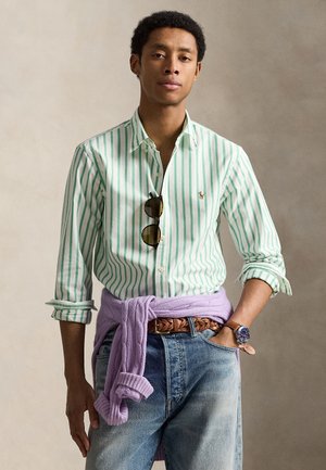 Moški, oblečen v zeleno-belo črtasto srajco z gumbi, sončna očala, obešena na ovratniku, sivkasto pulover, vezan okoli pasu, in jeans hlače.