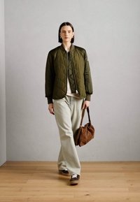 Quiltet olivenfarget bomberjakke, lys beige topp, løse lyse bukser, bærende en brun slouchy håndveske, med hvite og brune sko.