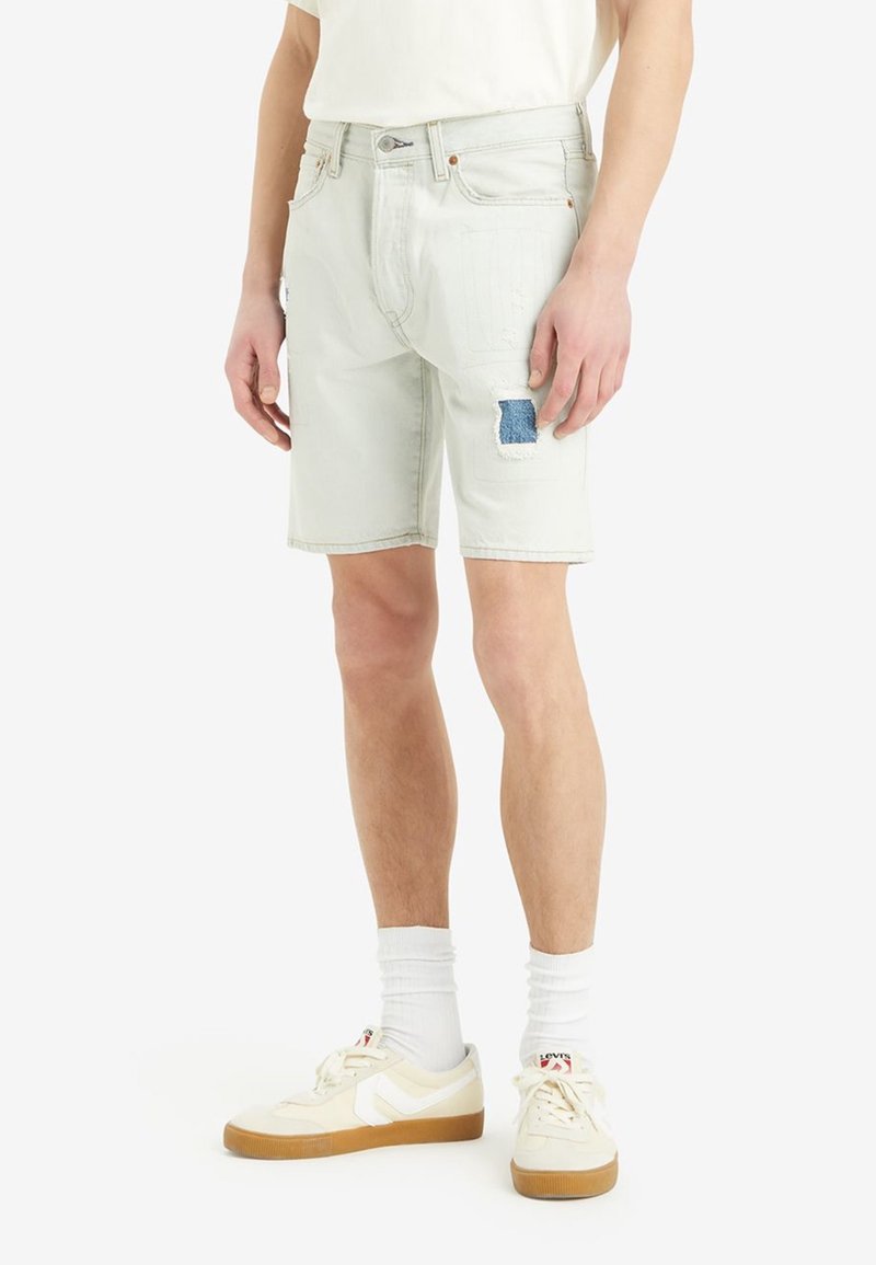 Homme portant un short en denim beige clair avec un patch bleu sur la cuisse gauche, des chaussettes blanches et des baskets beiges avec des semelles en gomme.