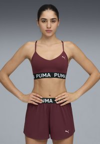 Bordo sportski grudnjak s tankim trakama, s crnim pojasom na kojem piše "PUMA" bijelim slovima, u kombinaciji s odgovarajućim bordom šortsevima i malim logotipom.