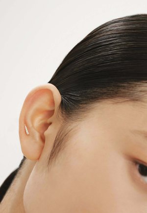 Pendiente de oro con piedras claras brillantes, que presenta un diseño angulado y elegante. Colocado en una oreja, con cabello liso visible cerca.