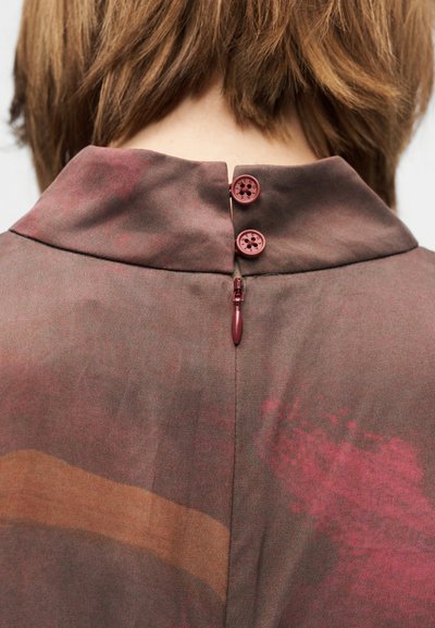 Rudos spalvos marškinėliai su apykakle, gale turintys du burgundiškos spalvos mygtukus ir užtrauktuko detales, pagaminti iš šiek tiek tekstūruotos medžiagos.